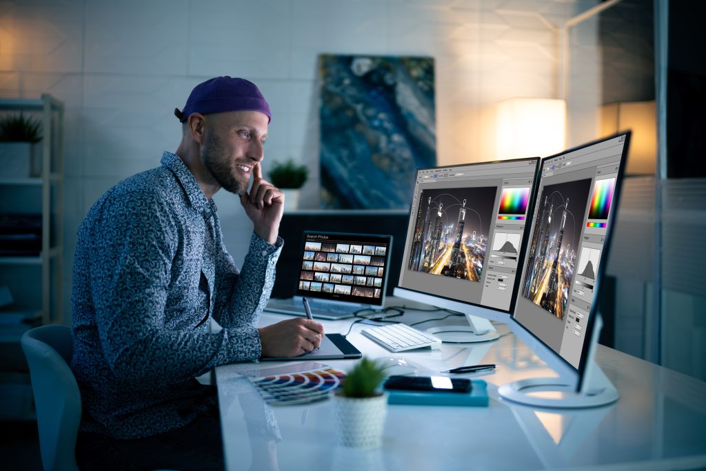 A man wearing a purple beanie and patterned shirt sits at a desk working on a digital art piece for his Book Cover Design Service displayed on dual monitors. The workspace is modern, with a small plant, tablet, color samples, and a lamp, creating a cozy, well-lit environment. A photograph is on the screen of a nearby laptop. BookSelf Book Cover Design & Premade Book Covers
