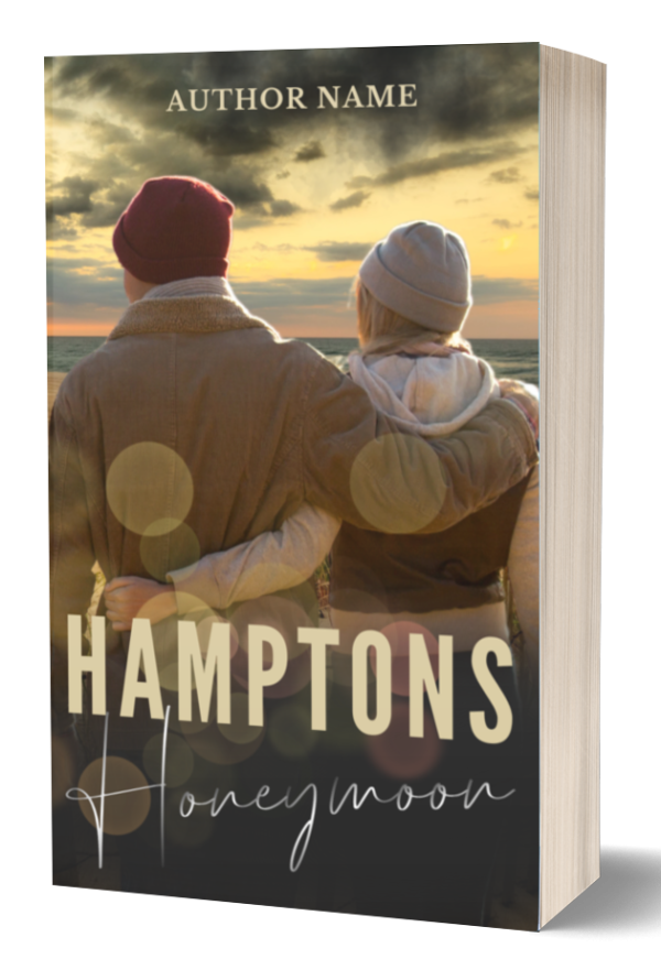 A paperback book titled "Hamptons Honeymoon: Ready Made Book Cover" with "Author Name" written above. The cover image features a couple standing arm in arm at a beach, watching the sunset over the ocean. They are dressed warmly in jackets and hats. The title is in large, bold letters, with "Honeymoon" in cursive. BookSelf Book Cover Design & Premade Book Covers
