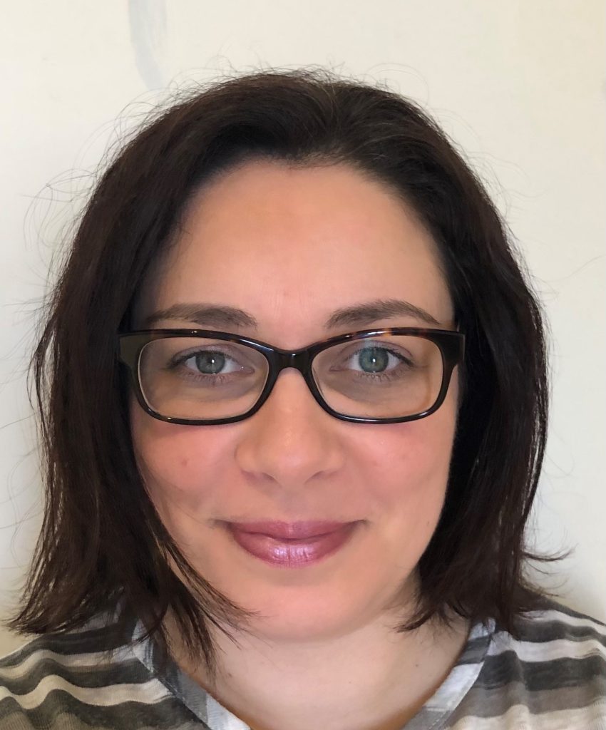A woman with shoulder-length dark brown hair is smiling at the camera, showcasing her profile. She is dressed in a gray and white striped top, wearing black-framed glasses and light pink lip gloss. The background is a plain white wall, highlighting her as though she were a poised designer. BookSelf Book Cover Design & Premade Book Covers