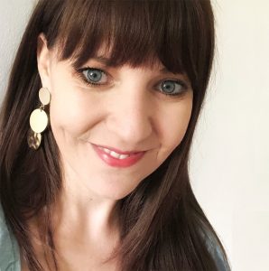 A woman with long brown hair and straight bangs smiles at the camera. She is wearing dangling gold earrings with circular designs and has blue eyes, light skin, and a light-colored top. The plain white background makes her the focal point of this designer profile. BookSelf Book Cover Design & Premade Book Covers